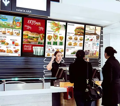 Scène dans un restaurant de restauration rapide, avec deux clients debout devant le comptoir en train de passer commande auprès d'un employé. Derrière le comptoir, des panneaux lumineux affichent le menu avec des images de différents plats et boissons.