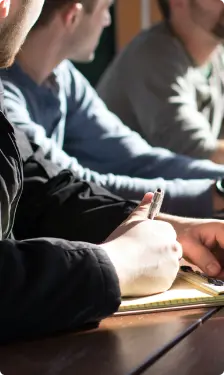 Groupe de personnes assises à une table, l'une d'elles prenant des notes sur un carnet. La scène suggère une réunion ou une séance de travail collaboratif.