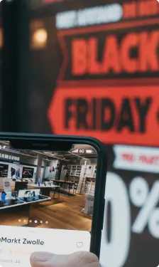 Une main tient un smartphone affichant une boutique, avec en arrière-plan une grande affiche publicitaire pour le Black Friday, mettant en avant des réductions.